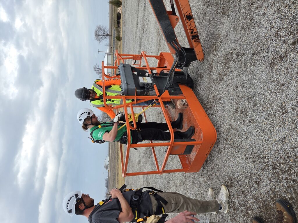 Three construction workers in hard hats and safety vests stand on an orange aerial lift, inspecting a control panel at a rural work site on a gravel lot.