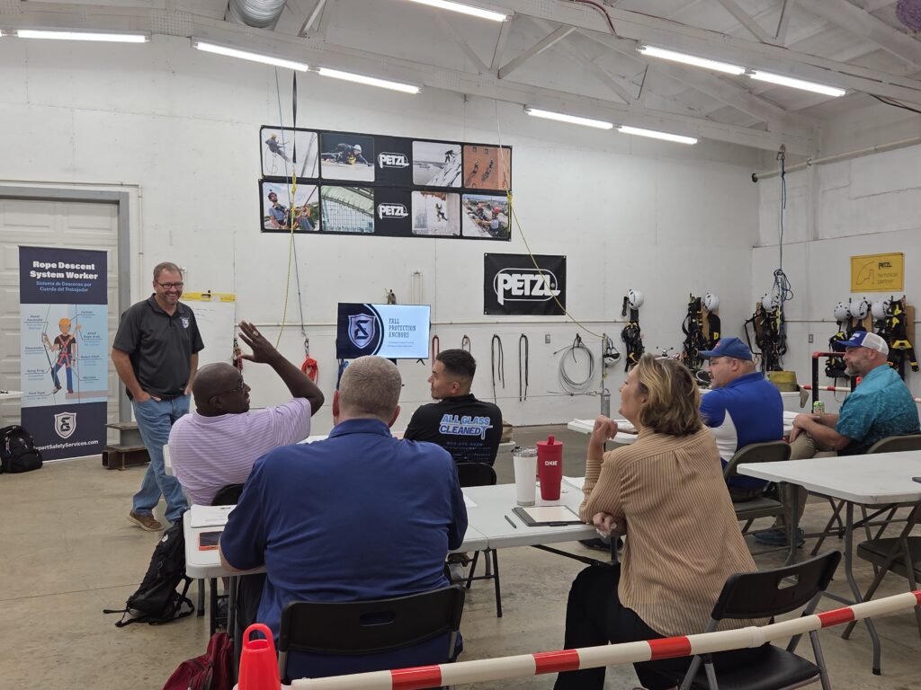 Group of people seated at tables in a climbing safety training workshop, instructor standing at front.
