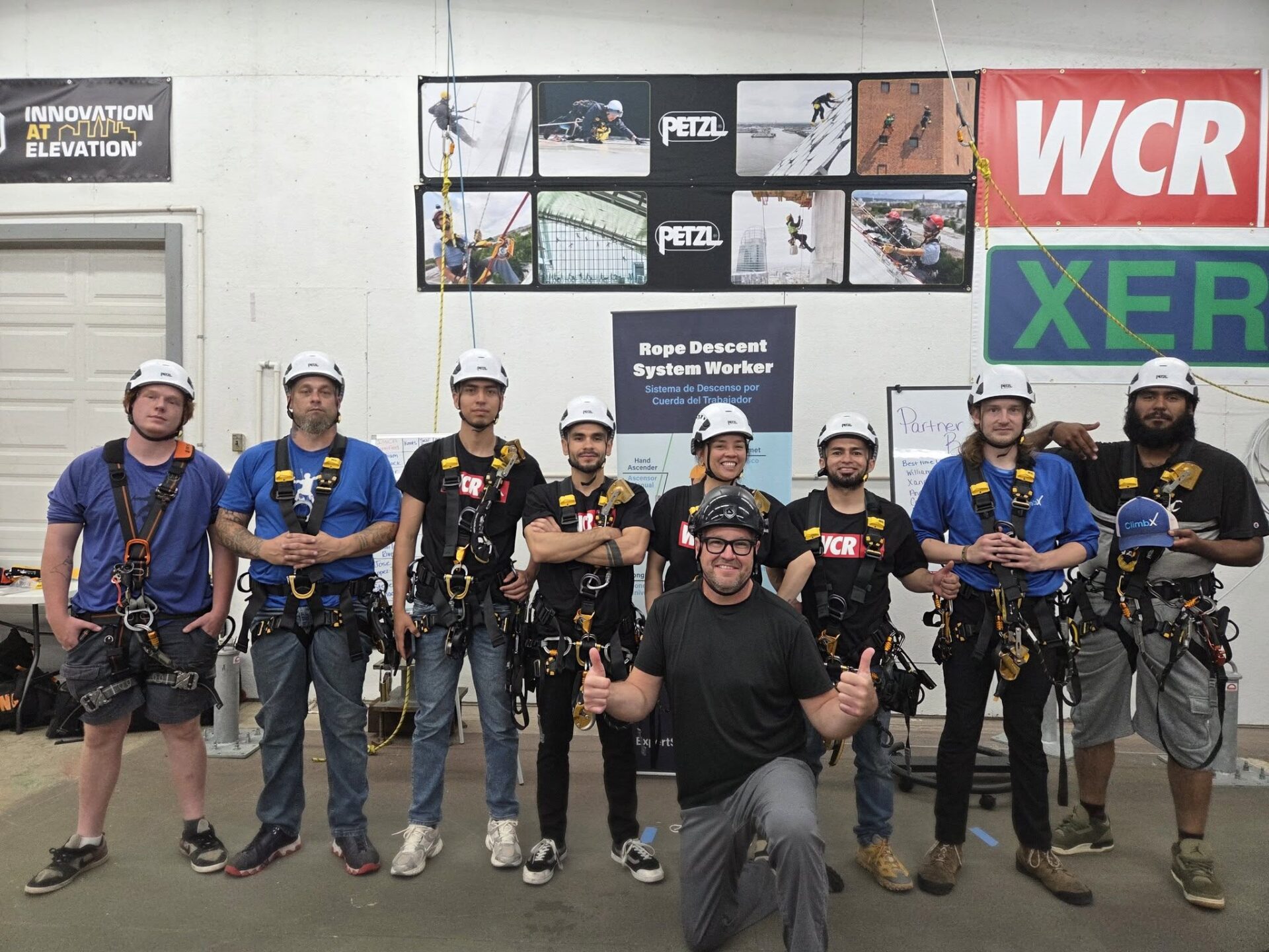 Group of climbers wearing helmets and safety harnesses posing indoors in front of banners and signage.